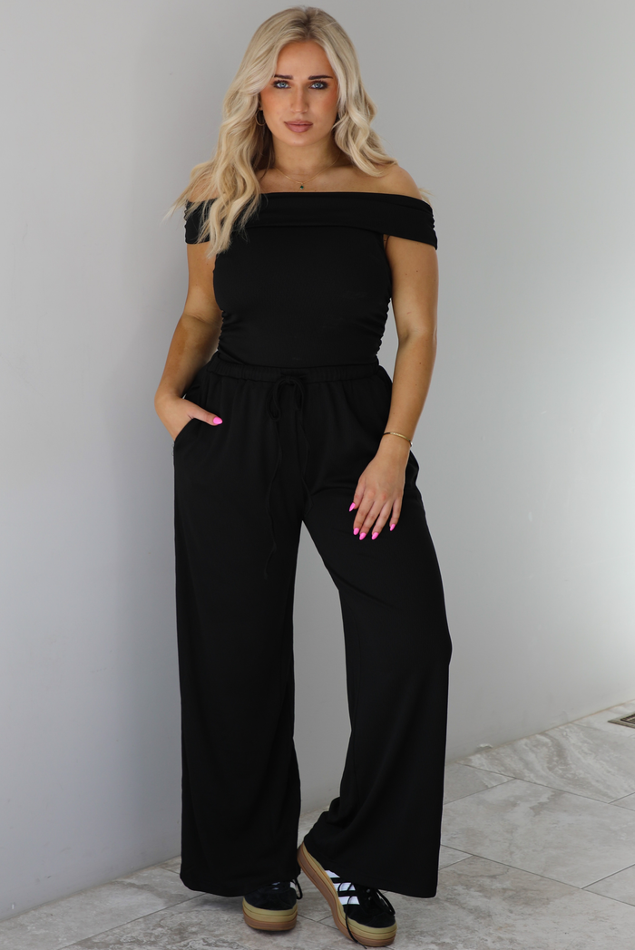 Woman wearing a two piece set featuring an off the shoulder top and high waisted pants. The photo is taken indoors against a white wall with white marble flooring. 