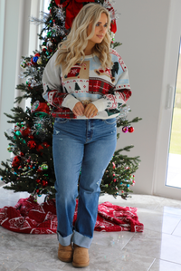 Woman model wearing a multi-colored and multi-patterned Christmas sweater with medium wash denim jeans. The photo is taken inside a modern home with a decorated Christmas tree in the back. 