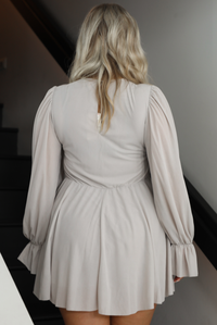 Woman model wearing a light taupe romper that looks like a dress. The photo is taken indoors against a white wall with a black railing and black steps. The photo shows the back that has a keyhole closure at the top and a cinched waistline. 