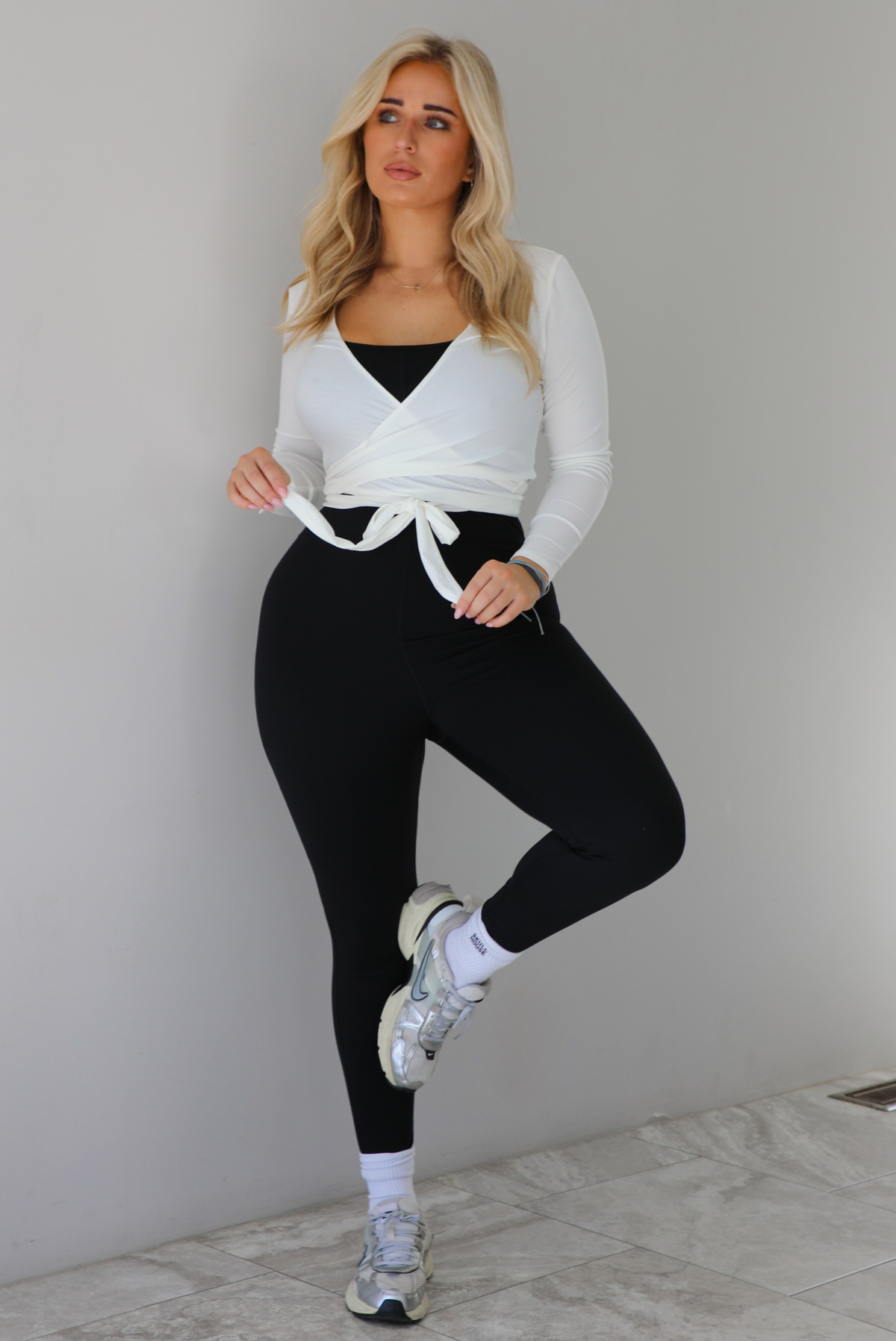Woman wearing a white cropped wrap style fitted top over a black fitted jumpsuit. The outfit is paired with white ankle socks and silver sneakers. The photo is taken indoors against a white wall with white marble flooring. 