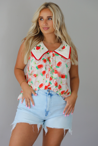 Woman wearing a cream and red strawberry patterned top with a collared detail and functional button closures in the front with light wash denim shorts indoors against a white wall. The photo shows the top tucked into the front of the shorts. 