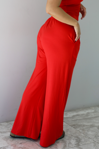 Woman wearing a red two piece set featuring a  tank top and slouchy pants. The photo shows a closeup of the back of the pants.The photo is taken indoors against a white wall with white marble flooring. 