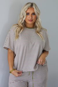 A woman wearing a matching short-sleeve striped lounge set in beige and olive green, featuring a crewneck top and drawstring pants, posed against a solid white wall. 
