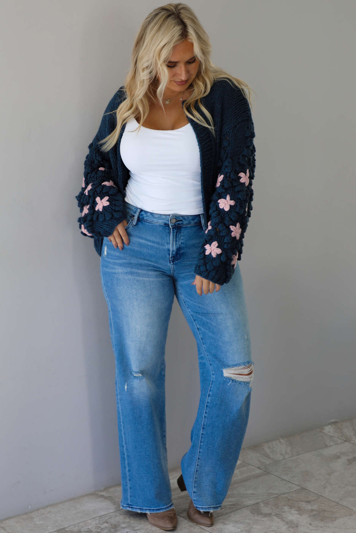 A woman wearing a navy oversized cardigan with a pink embroidered floral design on the sleeves, paired with a white tank, mid-rise medium wash denim jeans, standing indoors against a white wall with white marble flooring. 