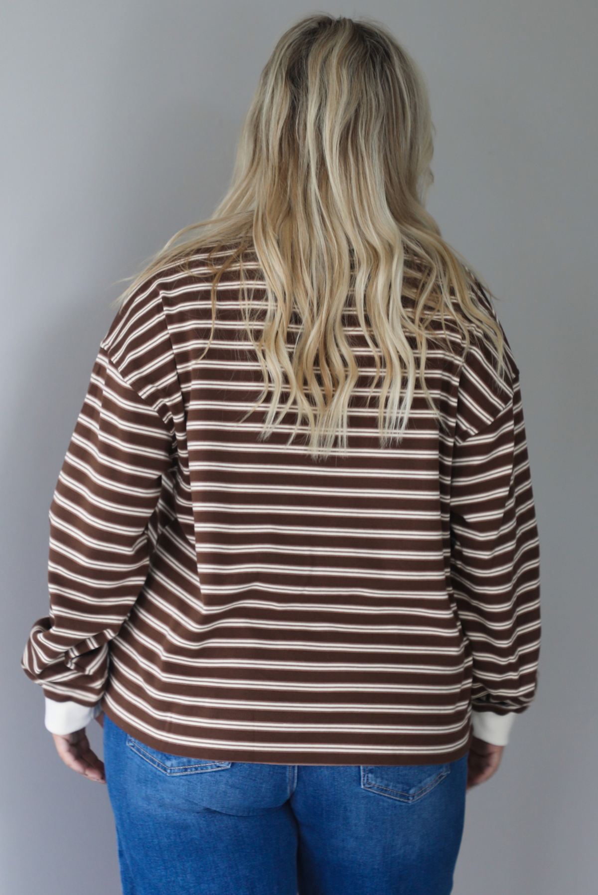 A woman shown from behind wearing a brown and cream striped collared quarter-button top, paired with dark, wide leg denim with a cuffed hem, standing indoors against a white wall. The top is untucked to show the full length. 