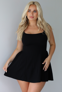 Woman wearing a black mini dress with thick straps, a fitted bodice, and flowy skirt indoors against a white wall.