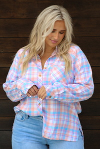 A woman wearing a pink, blue, and purple plaid print pattern throughout that has a collared neckline, functional button closures along the front, and an oversized fit, with one side tucked into light wash denim, standing in front of a dark brown wooden door. 