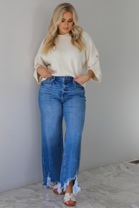 Woman wearing a cream colored quarter sleeve sweater that has ribbed details at the neckline, on the sleeves, and on the hem, with non-functional button closures on the sleeves. The sweater has a relaxed fit and is paired with a medium wash, high waisted, distressed hem denim with sand colored sandals. The photo is taken indoors against a white wall with white marble flooring. 