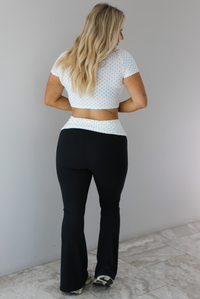 Woman wearing a two piece set featuring a white cropped top with a black polka dot print pattern and black pants that are high waisted with a white and black polka dot print folded over detail. The photo is taken indoors against a white wall with white marble flooring. The photo shows the back of the set. 