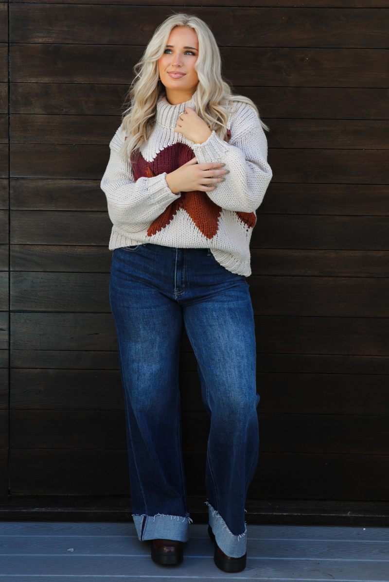 Woman model wearing a chunky knit turtle neck sweater with dark denim jeans outdoors in front of a dark wood door. 