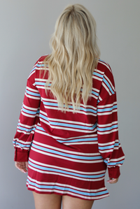 A woman shown from behind wearing a red burgundy mini dress with a white and blue striped pattern throughout, collared neckline, long sleeves, and functional pockets on ether side, standing indoors against a white wall.