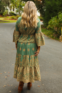 Woman wearing a teal dress that has tan a stitched pattern throughout, quarter length  sleeves, a collared, v-neckline, cinched with elastic waistline, and a maxi-length skirt. The photo shows the back of the dress, and is taken outdoors standing on a paved road with trees and other greenery in the background.