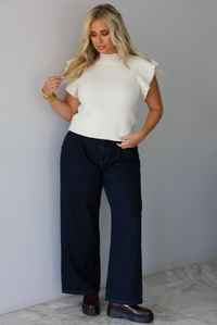 A woman wearing a cream ribbed knit sweater top with ruffled sleeves, dark wide-leg denim jeans, and brown loafers, standing indoors against a white wall with white marble flooring. 