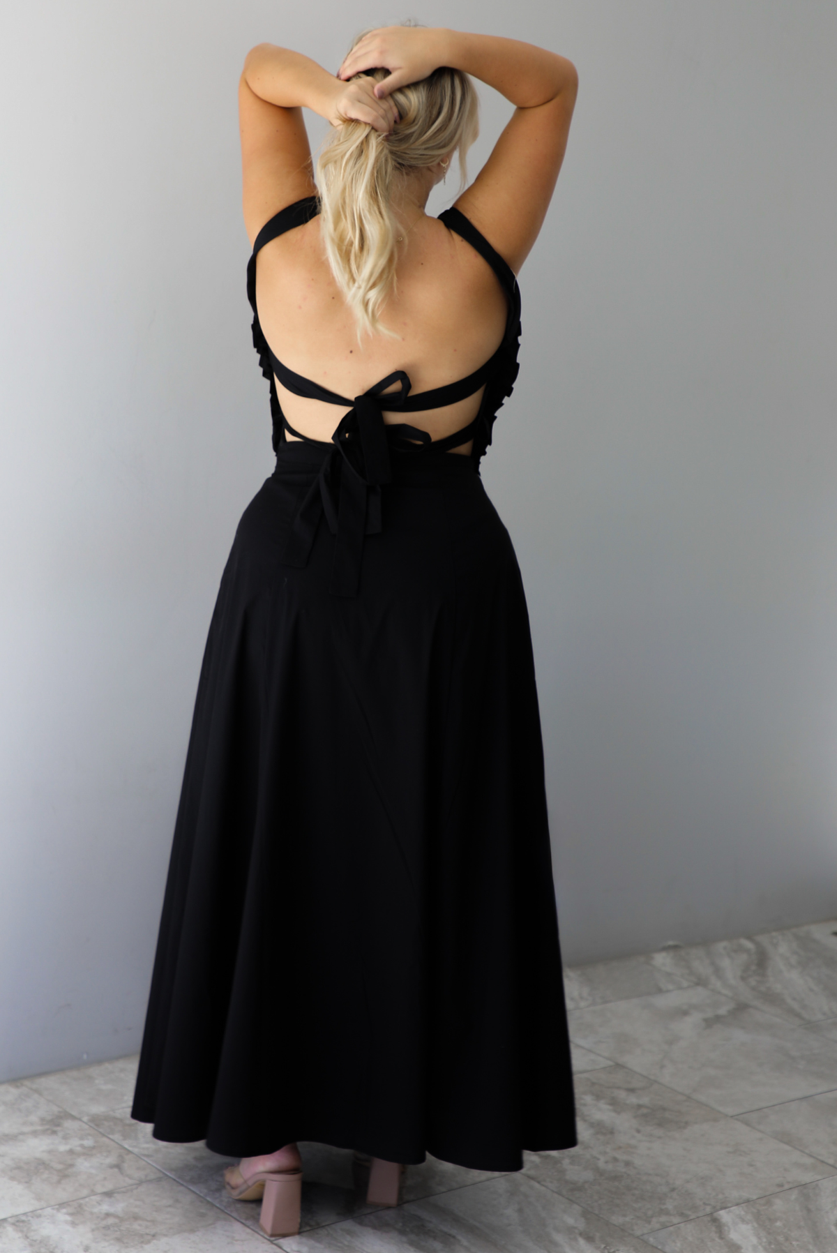 A woman shown from behind wearing a black two piece set featuring a backless top with tie closures and a high waisted maxi skirt, standing indoors against a white wall with white marble flooring. 