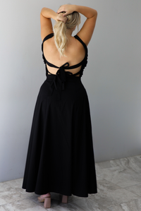 A woman shown from behind wearing a black two piece set featuring a backless top with tie closures and a high waisted maxi skirt, standing indoors against a white wall with white marble flooring. 