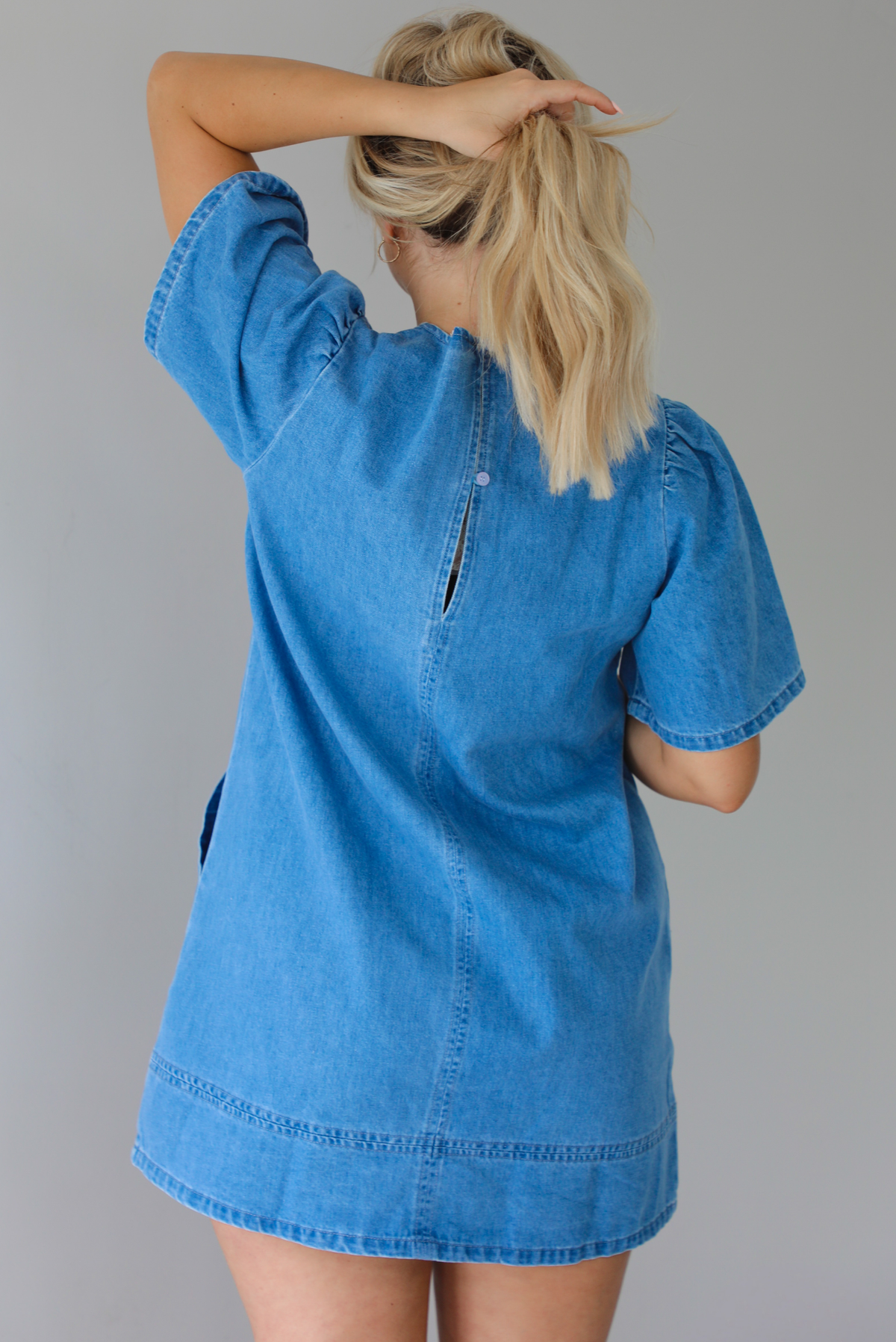 Woman wearing a v-neck, short sleeved, denim mini dress with functional pockets on either side. The photo is taken indoors against a white wall and shows the back of the dress. 