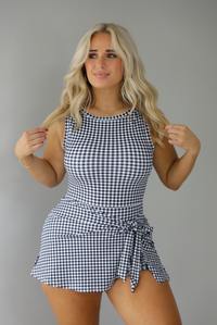 Woman wearing a black and white gingham print athletic romper that has a wrap-style skirt detail. The photo is taken indoors against a white wall.