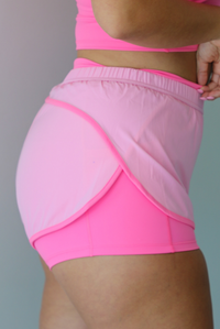 A woman modeling a bright pink two-piece activewear set consisting of a scoop-neck crop top and matching athletic shorts with a darker, hot pink trim, standing indoors against a white wall. The photo shows a closeup o the side of the shorts, displaying the fitted biker shorts underneath the looser light pink overlay. 