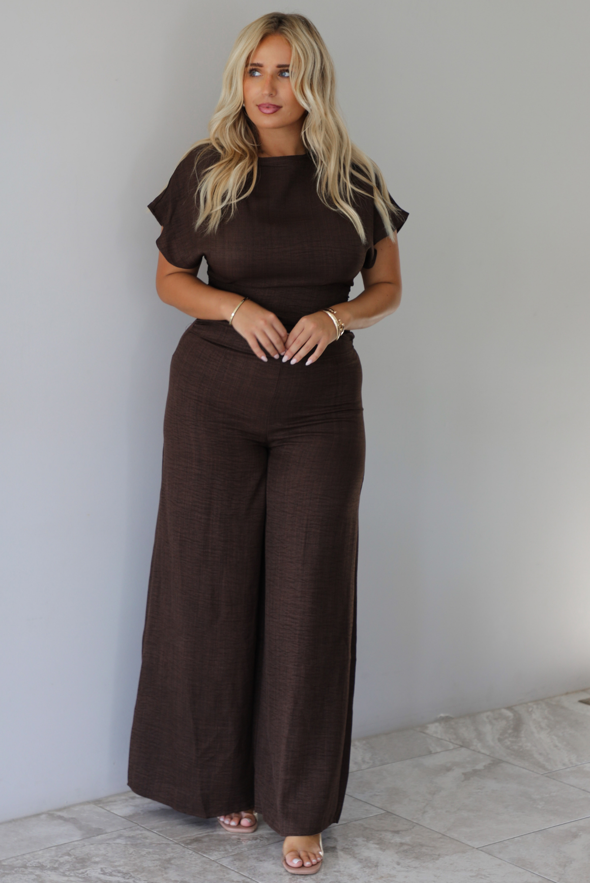 A woman modeling a dark brown short-sleeve jumpsuit with a high neckline and wide-leg pants, standing indoors against a white wall with white marble flooring. 
