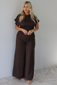 A woman modeling a dark brown short-sleeve jumpsuit with a high neckline and wide-leg pants, standing indoors against a white wall with white marble flooring. 