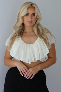 Woman wearing a cream ruffled cropped top with high waisted black, textured capri pants indoors against a white wall. The photo shows a closeup of the top.
