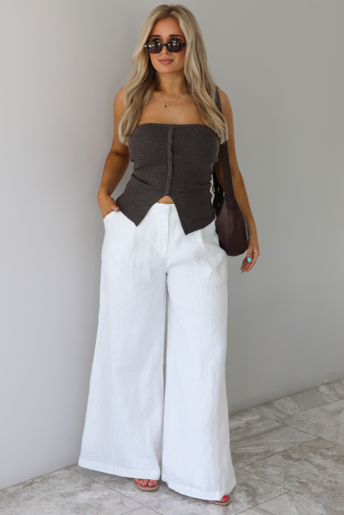 Woman wearing a strapless brown top with non-functional button closures in the front with  a split hem detail, and high waisted, flared hem white linen pants with a brown shoulder bag. The photo is taken indoors against a white wall with white marble flooring. 