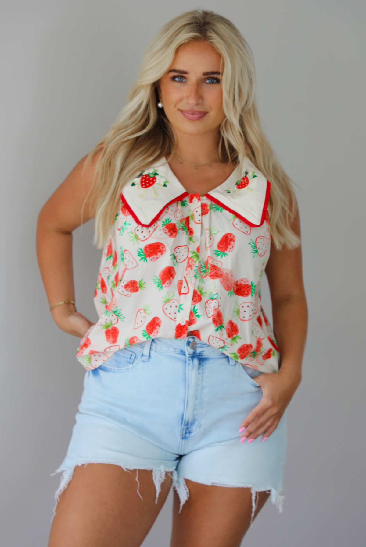 Woman wearing a cream and red strawberry patterned top with a collared detail and functional button closures in the front with light wash denim shorts indoors against a white wall. The photo shows the top tucked into the front of the shorts. 