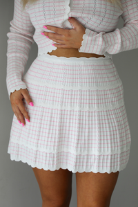 Woman wearing a two piece set that featuring a cropped cardigan and a high waisted skirt. The set is white with a pink horizontal striped pattern throughout, with scalloped hem details. The photo is taken indoors against a white wall. The photo shows a closeup of the skirt.