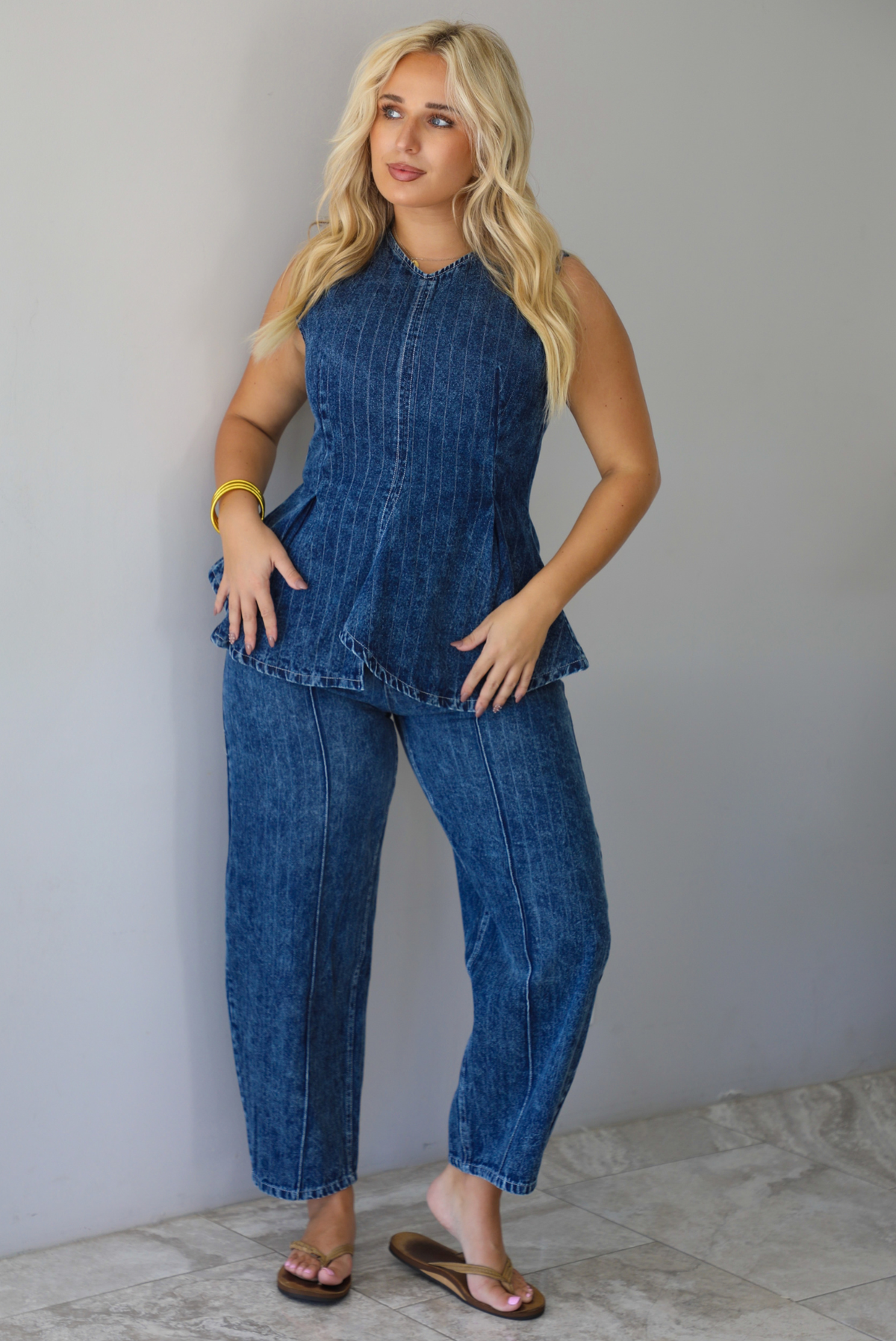 A woman wearing a matching two piece dark wash denim set with a pin striped design on it featuring a sleeveless peplum style top and high waisted denim jeans, standing indoors against a white wall with white marble flooring. 