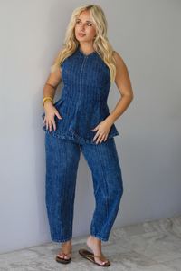 A woman wearing a matching two piece dark wash denim set with a pin striped design on it featuring a sleeveless peplum style top and high waisted denim jeans, standing indoors against a white wall with white marble flooring. 