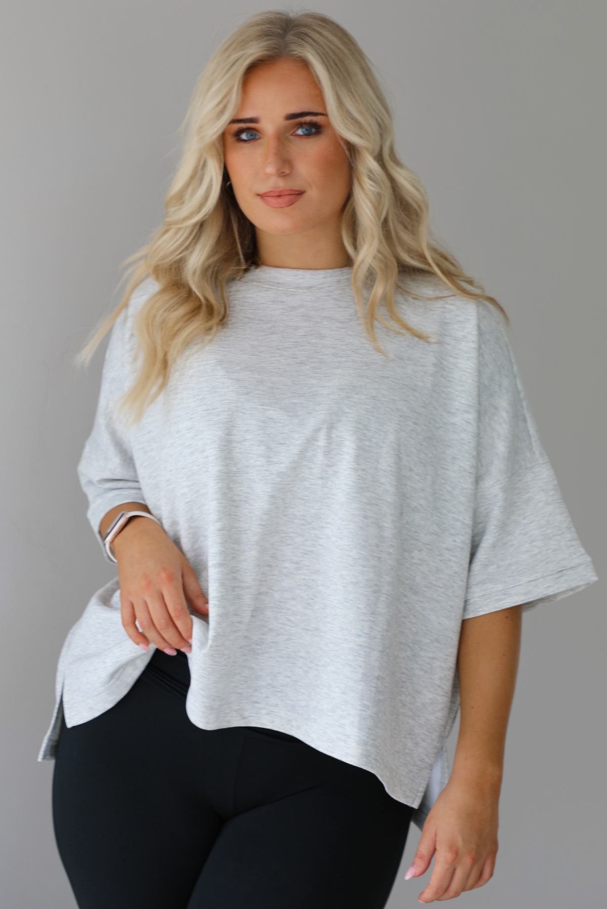 Woman wearing an oversized heather grey box tee with black leggings that flare slightly at the hems. The photo shows a closeup of the tee. The photo is taken indoors against a white wall.