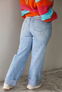 A woman shown from behind from the waist down wearing a pair of light wash denim jeans with a cuffed hem, standing indoors against a white wall with white marble flooring. 