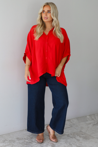 A woman wearing a bright red oversized short-sleeve blouse and dark wide-leg jeans, standing indoors against a white wall with white marble flooring. 