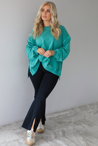 Woman wearing a teal oversized sweater with black leggings that have a slightly flared hem with split hem details in the front. The photo is taken in front of a white wall with white marble flooring. 