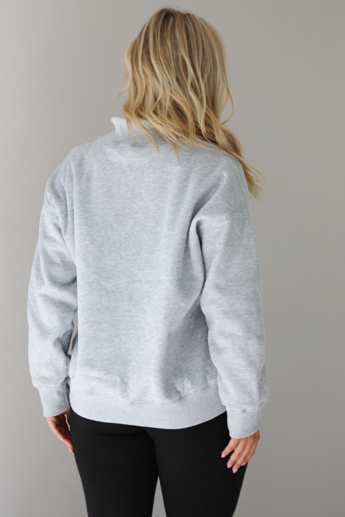 Woman wearing a grey turtleneck sweater, untucked, with high waisted black leggings. The photo shows the back of the sweater. The photo is taken indoors against a white wall.