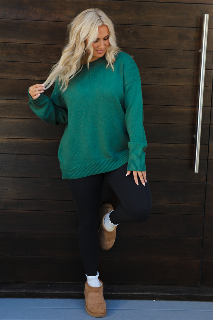 Woman model wearing a green oversized sweater with black leggings, white socks, and tan Uggs. The photo is taken outdoors against a dark brown door. 
