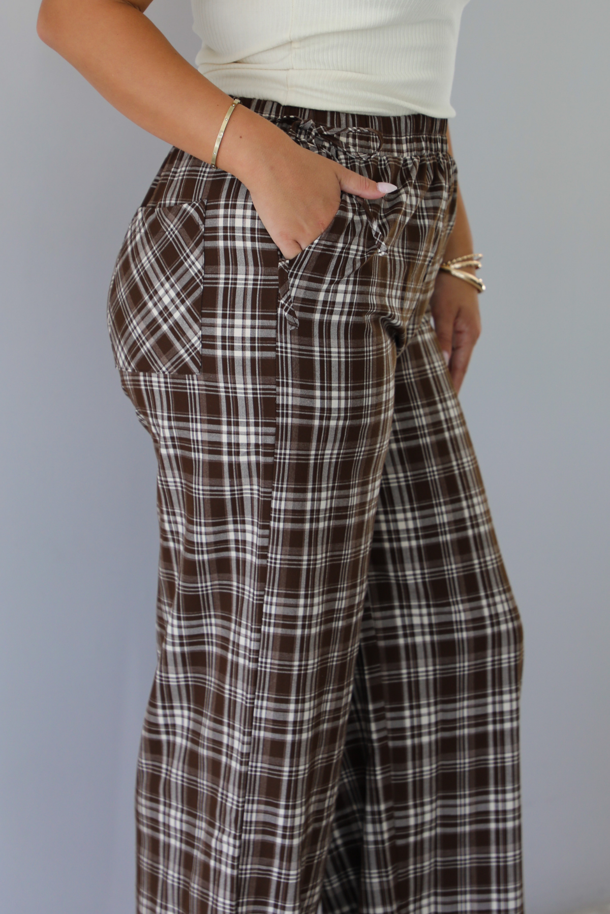 A person wearing brown, white, and light brown plaid wide-leg pants with a drawstring waistband, paired with a white ribbed top, shown from the side, focusing on the pockets, indoors against a white wall. 