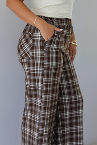 A person wearing brown, white, and light brown plaid wide-leg pants with a drawstring waistband, paired with a white ribbed top, shown from the side, focusing on the pockets, indoors against a white wall. 