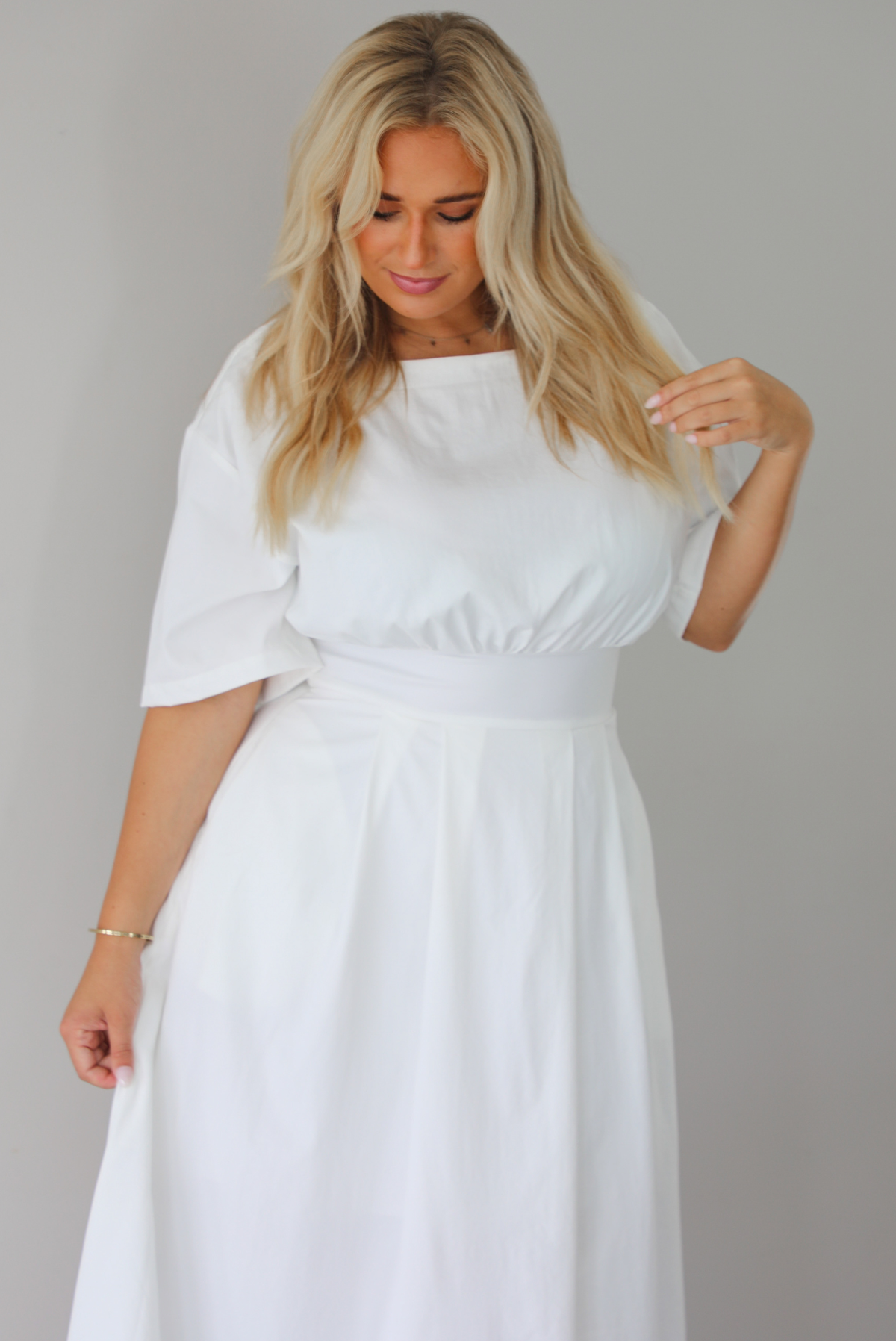 A woman modeling a white midi dress with short puff sleeves, a wide boat neckline, and side pockets, standing indoors against a white wall.