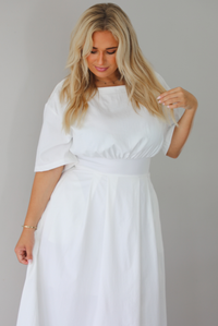 A woman modeling a white midi dress with short puff sleeves, a wide boat neckline, and side pockets, standing indoors against a white wall.