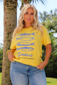 A woman wearing a bright yellow graphic t-shirt with blue sardine illustrations and the text 'Sardines LISBON FISH MARKET', paired with blue jeans, standing outdoors in front of a tree.