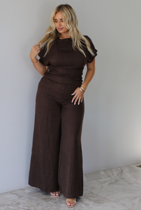 A woman modeling a dark brown short-sleeve jumpsuit with a high neckline and wide-leg pants, standing indoors against a white wall with white marble flooring. 