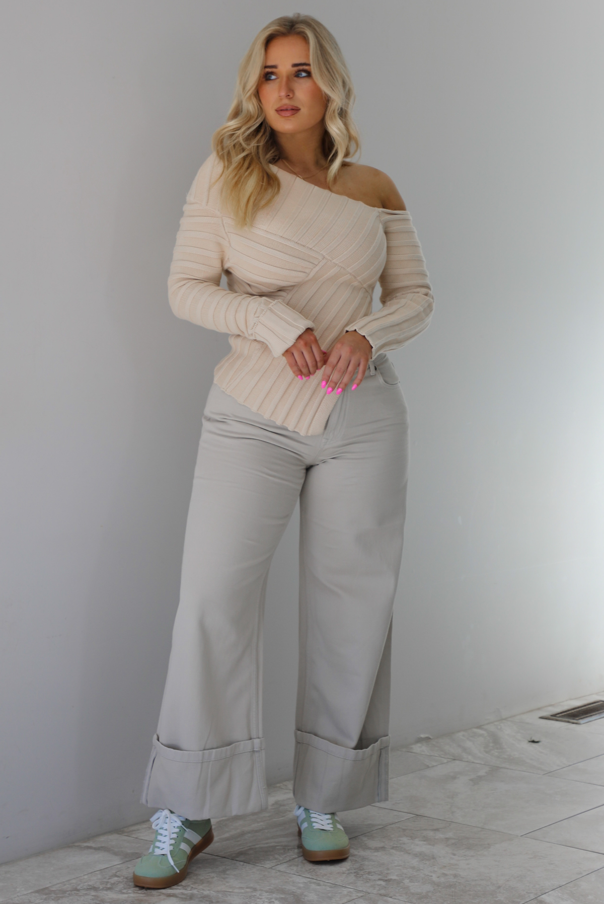 Woman wearing a cream colored ribbed textured knit top that has an asymmetrical hem with long sleeves in which one side is worn off the shoulder. The sweater is paired with sand colored high waisted, cuffed hem denim. The photo is taken indoors against a white wall with white marble flooring. 
