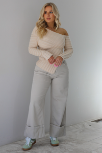 Woman wearing a cream colored ribbed textured knit top that has an asymmetrical hem with long sleeves in which one side is worn off the shoulder. The sweater is paired with sand colored high waisted, cuffed hem denim. The photo is taken indoors against a white wall with white marble flooring. 