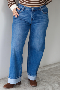 A closeup of a woman wearing dark wash, wide leg denim with a cuffed hem design, standing indoors against a white wall with white marble flooring. 