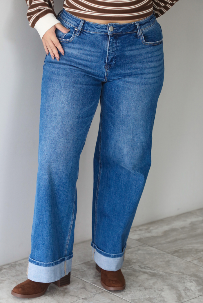 A closeup of a woman wearing dark wash, wide leg denim with a cuffed hem design, standing indoors against a white wall with white marble flooring. 
