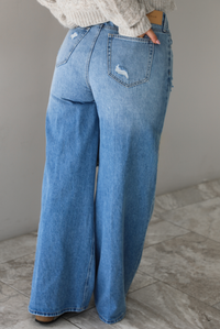 Woman wearing high waisted medium wash denim that have multiple button closures, functional belt loops, functional front and back pockets, and distressed details throughout with a flared hem. The photo shows the back of the denim. The photo is taken in front of a white wall and white marble flooring.