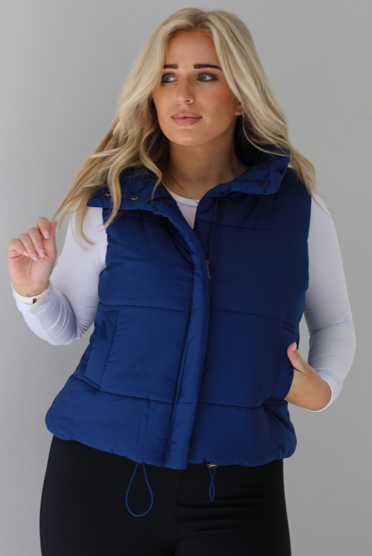 Woman wearing a navy puffer vest, white long-sleeve shirt, and black leggings indoors against a white wall. The puffer vest has a collared design, functional button closures at the neckline, a zipped zipper closure in the front, and functional pockets on either side.