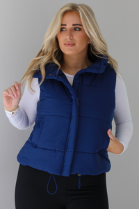 Woman wearing a navy puffer vest, white long-sleeve shirt, and black leggings indoors against a white wall. The puffer vest has a collared design, functional button closures at the neckline, a zipped zipper closure in the front, and functional pockets on either side.