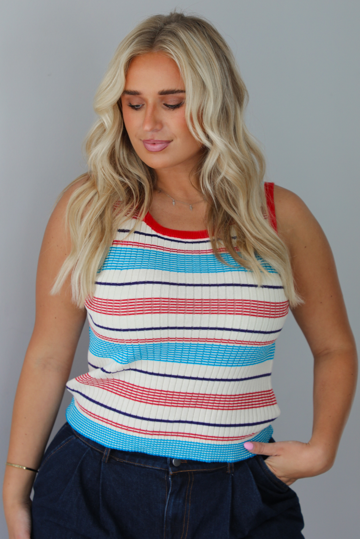 A woman modeling a sleeveless ribbed knit top with horizontal stripes in red, light blue, navy, and white, paired with dark wide-leg jeans and metallic rose gold flats, standing indoors against a white wall.